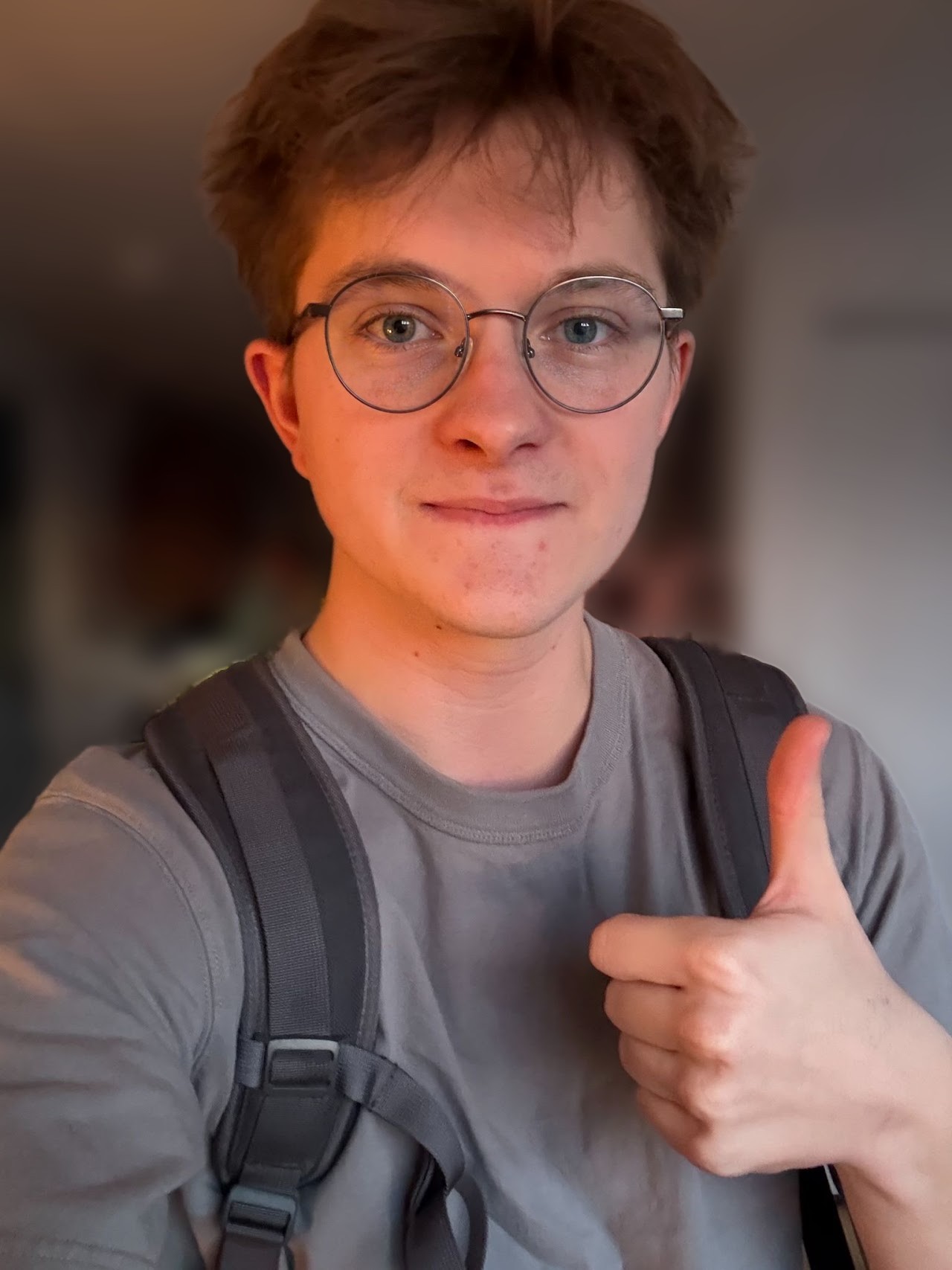 Max, wearing round glasses and a backpack, giving a thumbs up in a sunlit hallway